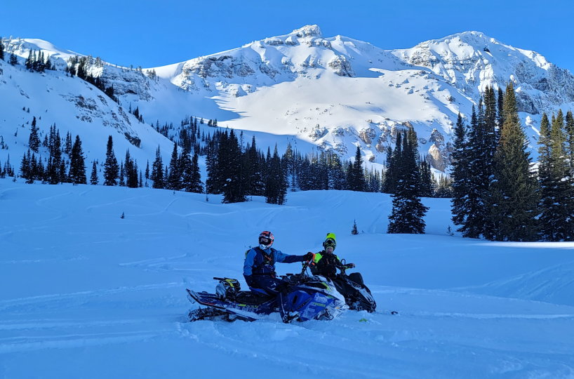 Two people riding a snowmobile in a snowy mountain landscape.